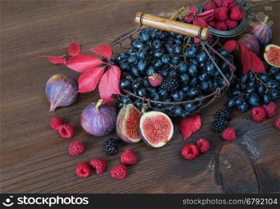 Black grapes in a basket and ripe figs, red raspberries and blackberries with autumn leaves on the wooden background