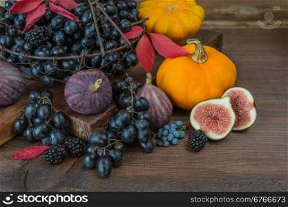 Black grapes in a basket and ripe figs, red raspberries, orange pumpkin and blackberries with autumn leaves on the wooden background
