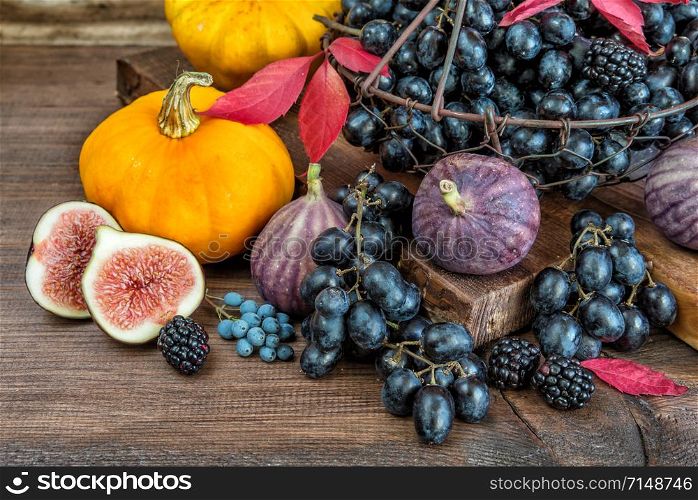 Black grapes in a basket and ripe figs, red raspberries, orange pumpkin and blackberries with autumn leaves on the wooden background