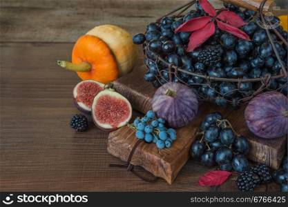 Black grapes in a basket on the old cutting board as well as ripe figs, red raspberries, orange pumpkin and blackberries with autumn leaves on the wooden background