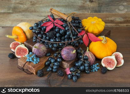 Black grapes in a vintage metal basket with wooden handle and ripe figs, red raspberries, orange pumpkin and blackberries with autumn leaves on the wooden background