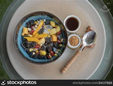 Black Grass Jelly is including Nuts, Cereals, Red beans, Ginkgo, Sliced   pumpkin, Corn kernels, Millet, Sweet potato, Palm seed and Jelly which is served with Longan juice and Brown sugar on Ceramic tray. Traditional iced sweet dessert, Top view, Space for text,  Selective focus.