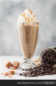 Black iced coffee with fresh milk and caramel with cream in classic milkshake glass and jar of raw coffee beans on light table background.