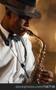 Black jazzman in hat plays the saxophone on the stage with spotlights. Black jazz musician preforming on the scene. Black jazzman in hat plays the saxophone on stage