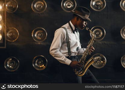 Black jazzman in hat plays the saxophone on the stage with spotlights. Black jazz musician preforming on the scene. Black jazzman in hat plays the saxophone on stage