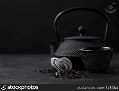 Black loose tea with strainer infuser and iron japanese teapot and cup on dark background.