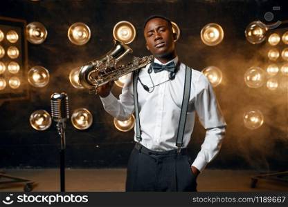 Black male jazz performer poses with saxophone on the stage with spotlights. Black jazzman preforming on the scene. Black jazz performer poses with saxophone on stage