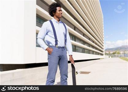 Black male worker with afro hairstyle standing next to an office building with a skateboard.. Black male worker standing next to an office building with a skateboard.
