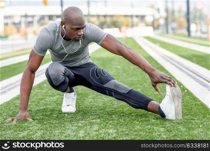 Black man doing stretching before running in urban background. Young male exercising listening to music with headphones.