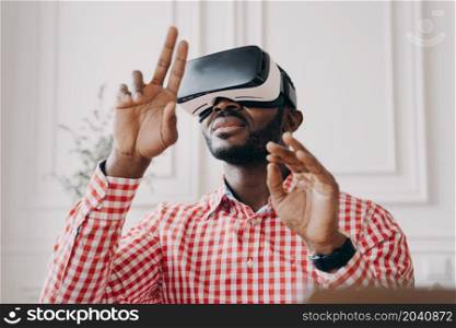 Black man in VR glasses experiencing 3D technology of augmented reality. Excited African male waving with hands up and down makes touching moves with fingers, stands against home office background. Black man in VR glasses experiencing 3D technology of augmented reality