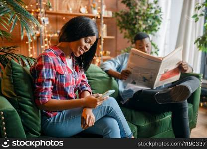Black man reading newspaper, his woman using mobile phone on couch, relaxation at home. Happy african love couple leisures in their house. Black man reading newspaper, his woman using phone