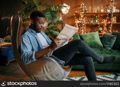 Black man reading newspaper in a comfortable leather chair in the living room, relaxation at home. Young african american male person in his house