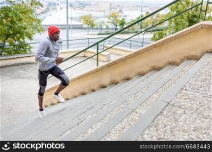 Black man running upstairs outdoors listening to music with white headphones. Young male exercising with city scape at the background.. Black man running upstairs outdoors in urban background