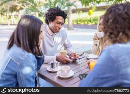 Black man showing something to his multi-ethnic group of friends on his smartphone, while having drinks at an outdoor table in a bar.. Black man showing his smartphone to his group of friends while having drinks at an outdoor bar