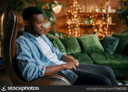 Black man sitting in a comfortable leather chair in the living room, relaxation at home. Young african american male person in his house