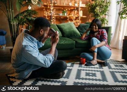 Black man takes photo, his woman poses at the couch in the living room, relaxation at home. Happy african love couple leisures in their house