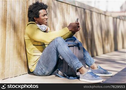 Black man with afro hair and headphones resting sitting on the ground using smartphone.. Black man using smartphone resting on the ground.