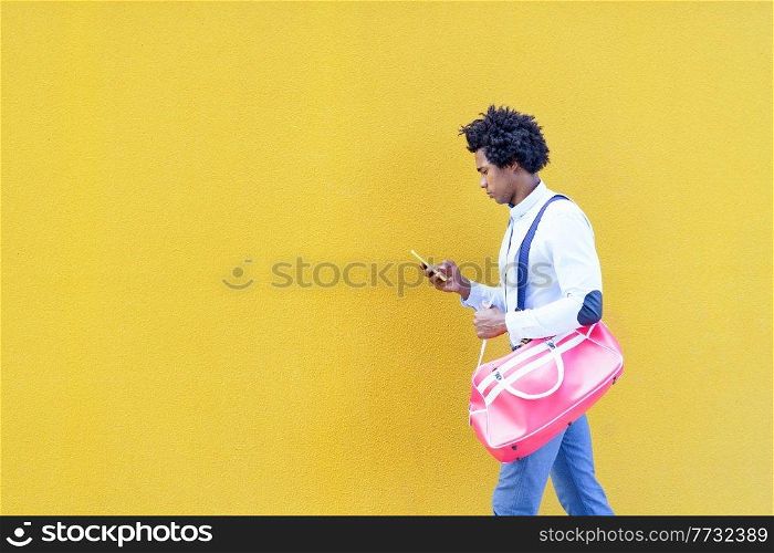Black man with afro hairstyle carrying a sports bag and smartphone against a yellow urban background. Guy with curly hair wearing shirt and suspenders.. Black man with afro hairstyle carrying a sports bag and smartphone in yellow background.