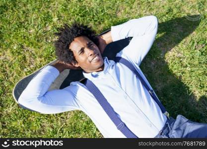 Black man worker with afro hair resting lying on the grass with his skateboard. Black businessman resting lying on the grass with his skateboard