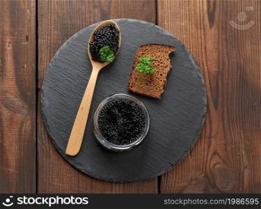 black paddlefish caviar in a wooden spoon, a slice of bread and a jar on a black round graphite board, top view