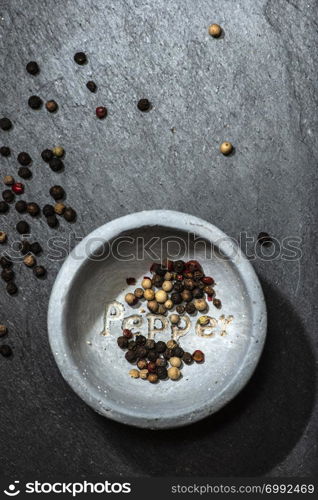 Black pepper in small bowl for spices on dark background. Red, green and black pepper grains close-up and natural light on it. Dark stone background and bowl with text Pepper on the bottom.