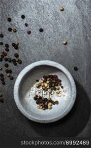 Black pepper in small bowl for spices on dark background. Red, green and black pepper grains close-up and natural light on it. Dark stone background and bowl with text Pepper on the bottom.