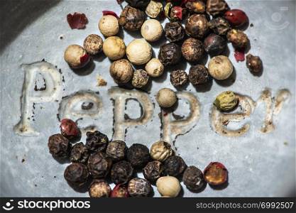Black pepper in small bowl for spices on dark background. Red, green and black pepper grains close-up and natural light on it. Dark stone background and bowl with text Pepper on the bottom.