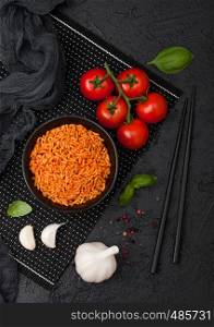 Black plate bowl of rice with tomato and basil and garlic and chopsticks on black background.