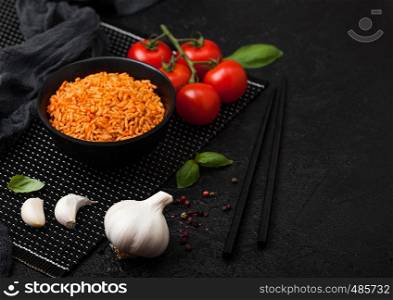 Black plate bowl of rice with tomato and basil and garlic and chopsticks on black background.