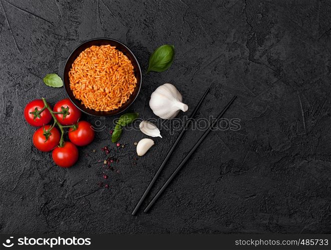 Black plate bowl of rice with tomato and basil and garlic and chopsticks on black background. Space for text