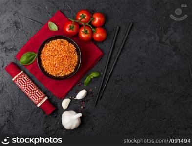 Black plate bowl of rice with tomato and basil and garlic and chopsticks on red napkin and bamboo place mat on black background. Top view. Space for text