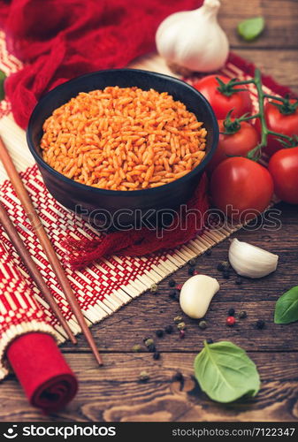 Black plate bowl of rice with tomato and basil and garlic and chopsticks on red bamboo place mat on wood background with red cloth. Top view.