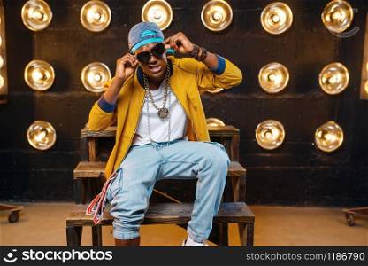 Black rapper in cap and sunglasses sitting on the steps, singer on stage with spotlights on background. Rap performer on scene with lights, underground music, urban style