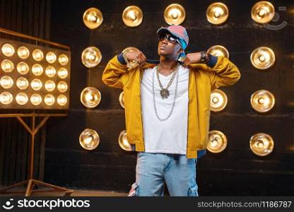 Black rapper in cap, perfomance on stage with spotlights on background. Rap performer on scene with lights, underground music, urban style. Black rapper in cap, perfomance on stage