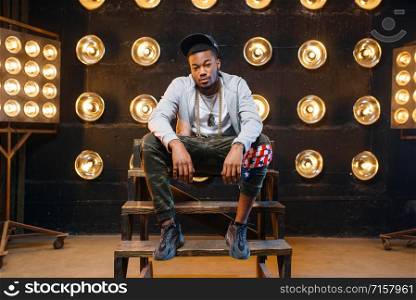 Black rapper in cap poses, perfomance on stage with spotlights on background. Rap performer on scene with lights, underground music. Black rapper in cap poses, perfomance on stage