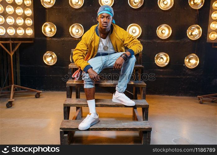 Black rapper in cap sitting on the steps, singer on stage with spotlights on background. Rap performer on scene with lights, underground music, urban style