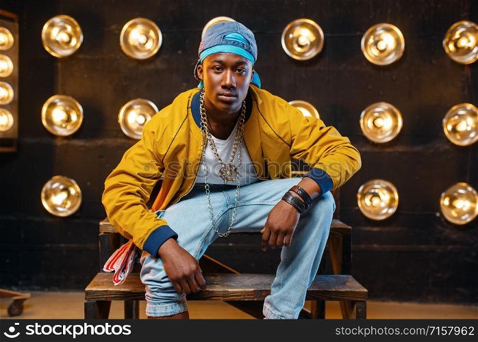 Black rapper in cap sitting on the steps, singer on stage with spotlights on background. Rap performer on scene with lights, underground music. Black rapper in cap sitting on the steps