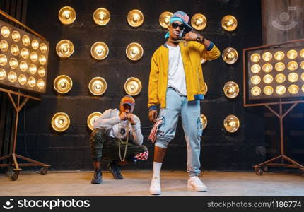 Black rappers in caps dance on stage with spotlights on background. Rap performers on scene with lights, underground music, urban style. Rappers in caps dance on stage with spotlights