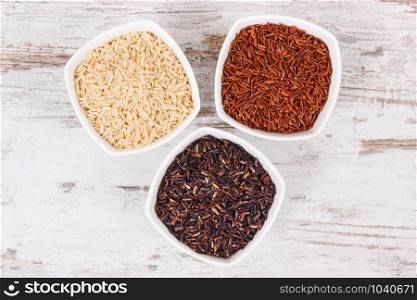 Black, red and brown rice in white glass bowls on rustic board, concept of healthy, gluten free food and nutrition. Black, red and brown rice in bowls, healthy gluten free food concept