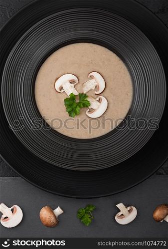 Black restaurant plate of creamy chestnut champignon mushroom soup on black background with black stone board and fresh mushrooms.