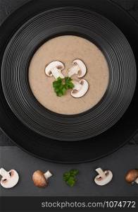Black restaurant plate of creamy chestnut champignon mushroom soup on black background with black stone board and fresh mushrooms.