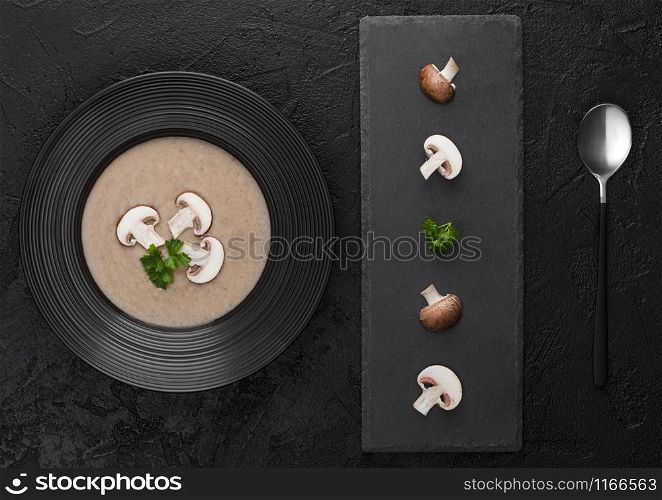 Black restaurant plate of creamy chestnut champignon mushroom soup on black background with black stone board and fresh mushrooms. Top view.