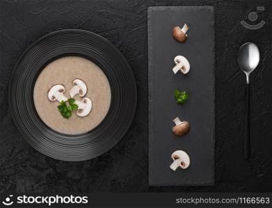 Black restaurant plate of creamy chestnut champignon mushroom soup on black background with black stone board and fresh mushrooms. Top view.