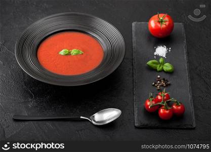Black restaurant plate of creamy tomato soup and spoon on black background with stone chopping board and raw tomatoes, pepper and salt.
