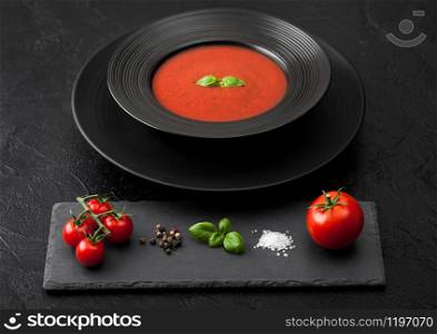 Black restaurant plate of creamy tomato soup on black background with stone chopping board and raw tomatoes, pepper and salt.