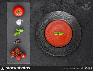 Black restaurant plate of creamy tomato soup on black table background with stone chopping board and raw tomatoes, pepper and salt. Top view