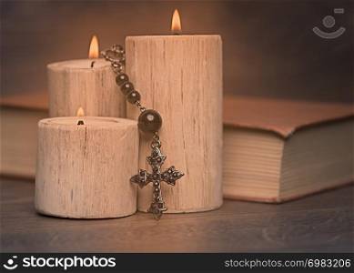black rosary and crucifix resting on closed book near the candles on wooden table, religion school concept. Vintage style.
