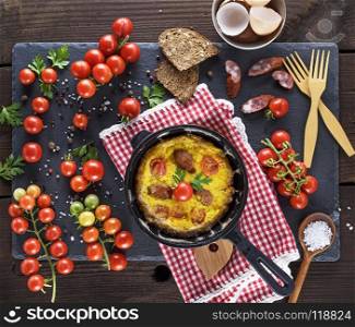 black round frying pan with fried omelette, next to fresh ripe red cherry tomatoes, top view
