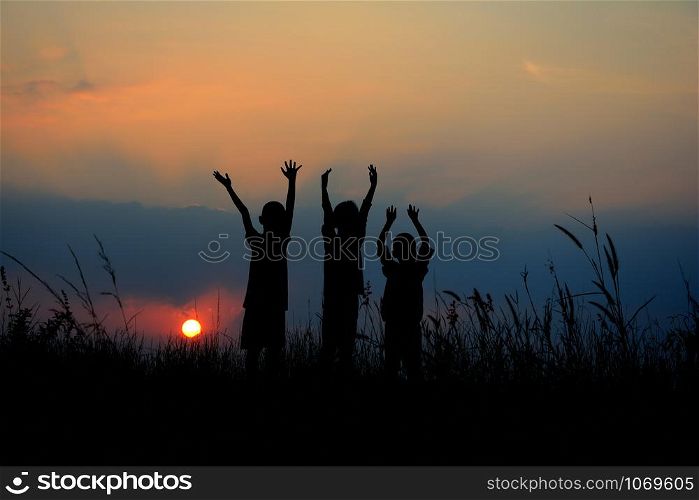 Black silhouette of three children standing together. There is a sky at sunset.