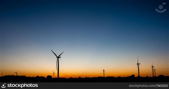 Black silhouettes of several wind turbines against orange and blue sky at dusk.